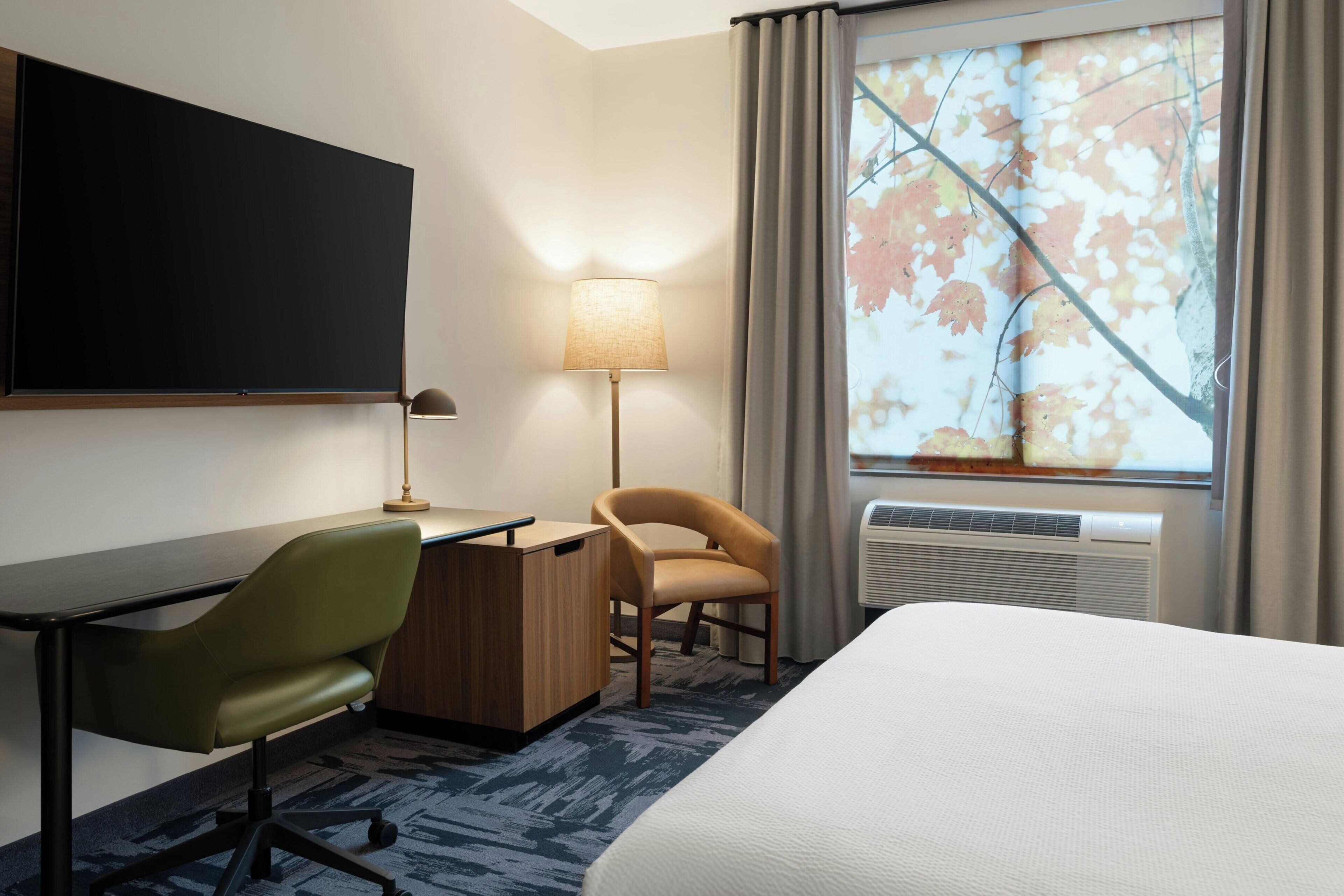 Habitación de hotel con cama, escritorio, silla, lámpara y un ventanal con cortinas.