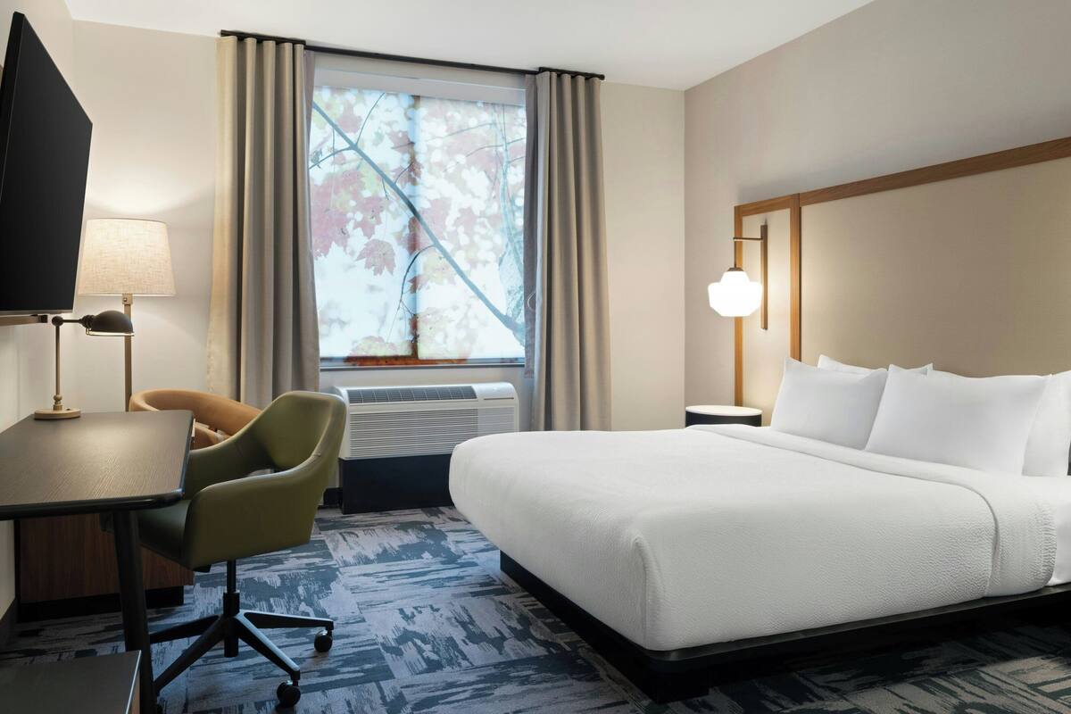 Quarto de hotel com cama, escrivaninha, cadeira, abajur, janela com cortinas e luminária na parede.