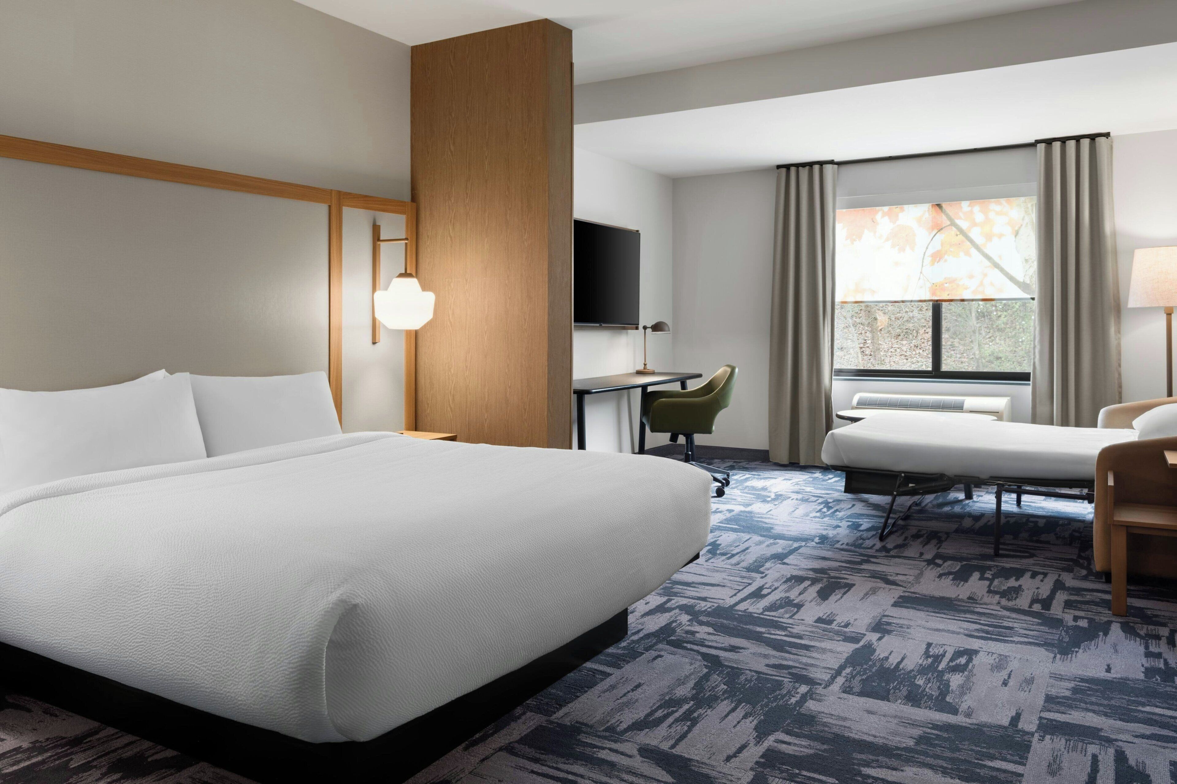 A hotel room with a large bed, a desk, a chair, a TV, and a window with curtains.