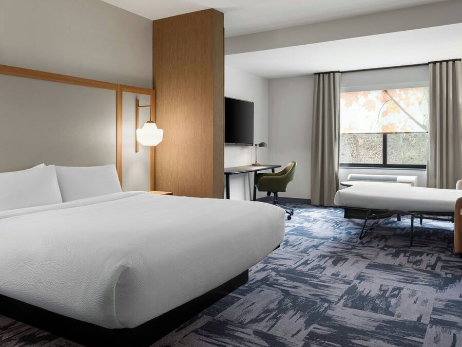 A hotel room with a large bed, a desk, a chair, a TV, and a window with curtains.