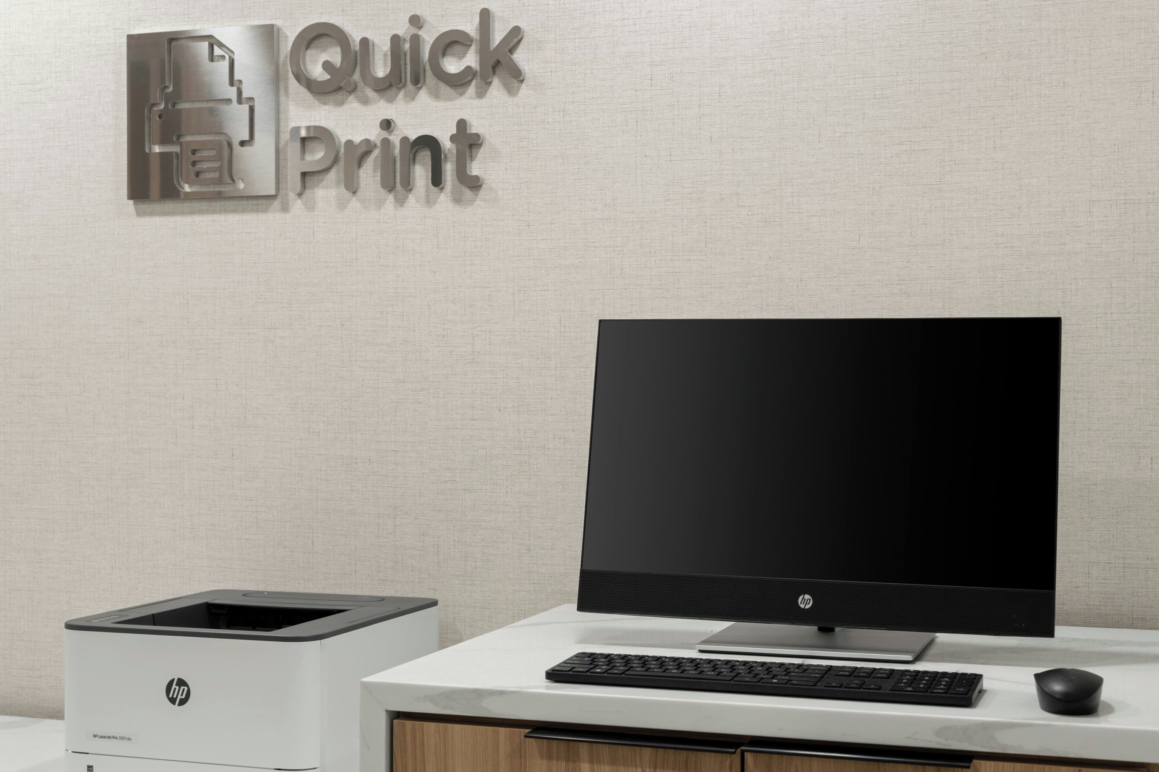 En HP Quick Print printer, en stationær computer med skærm, tastatur og mus på et hvidt skrivebord foran en væg med struktur.