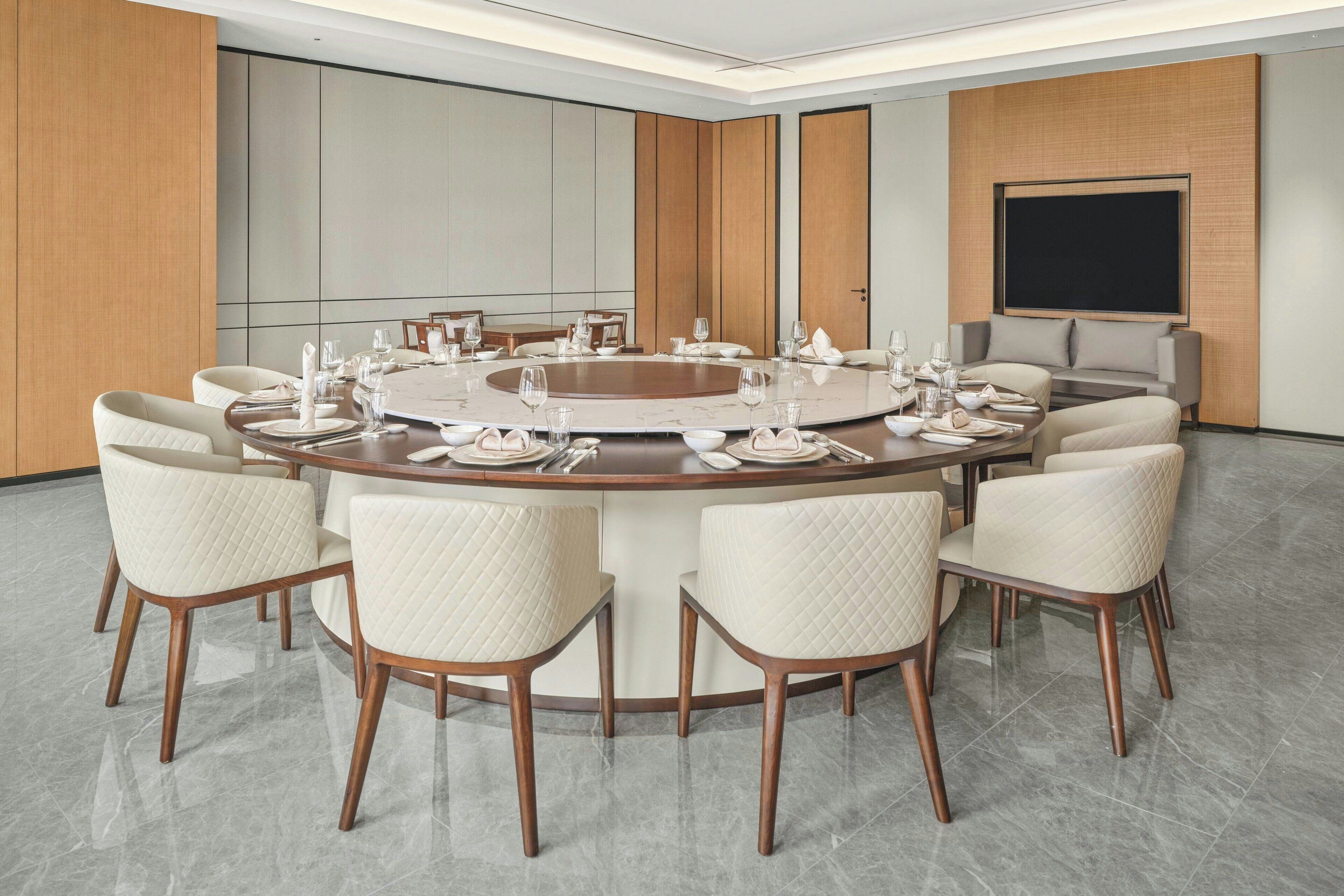 Un salón de conferencias moderno con una mesa redonda, sillas blancas y una pantalla grande.