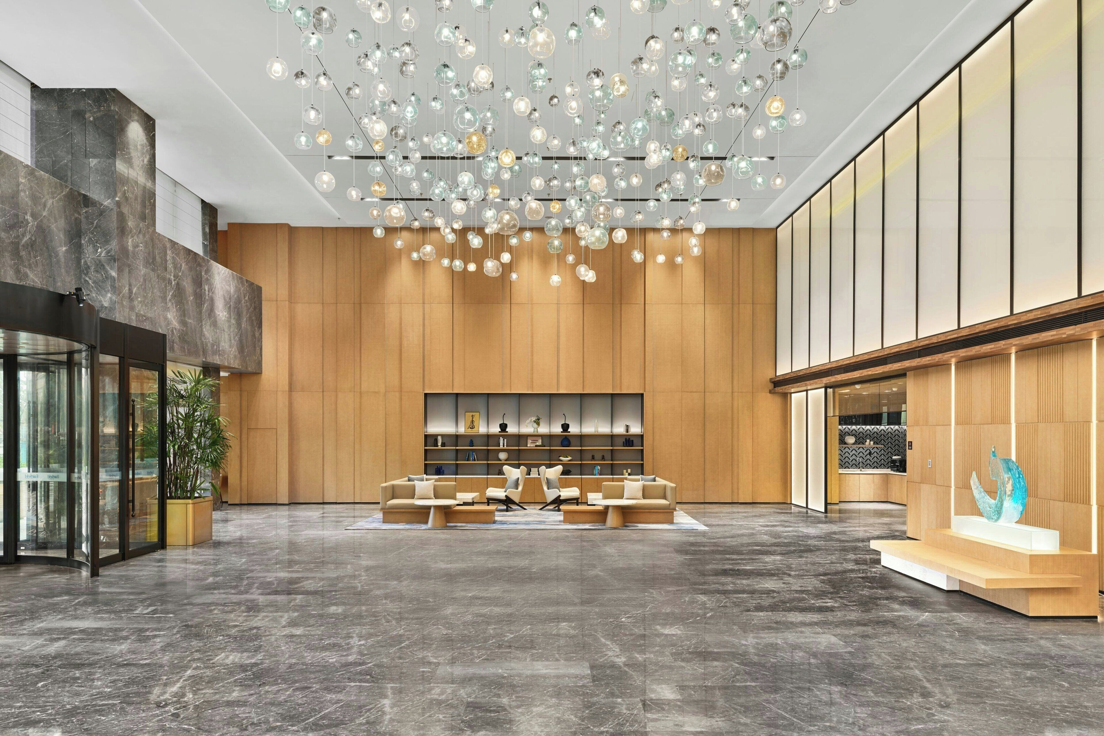 Un hall d’entrée spacieux au design moderne, comprenant un espace détente avec des canapés et des fauteuils, un grand mur recouvert de panneaux en bois et un luminaire au plafond au design original.