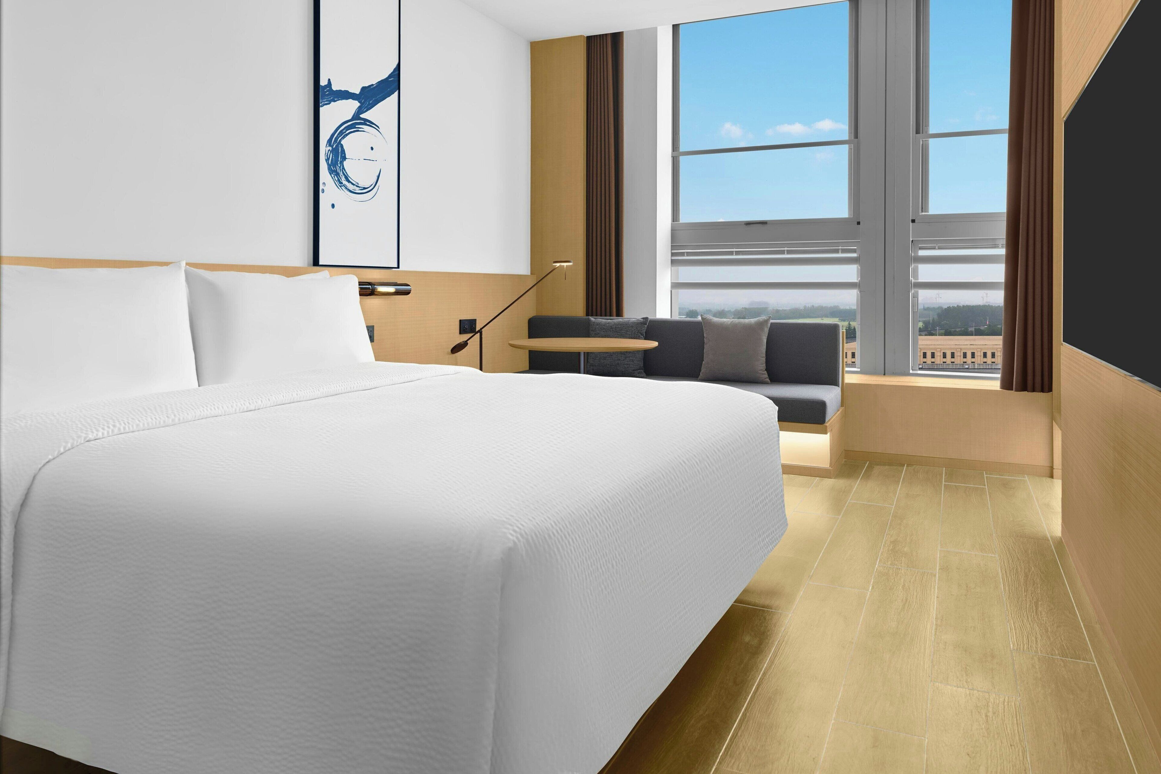 A modern hotel room with a large bed, a sofa, a small table, and a view of the city through large windows.