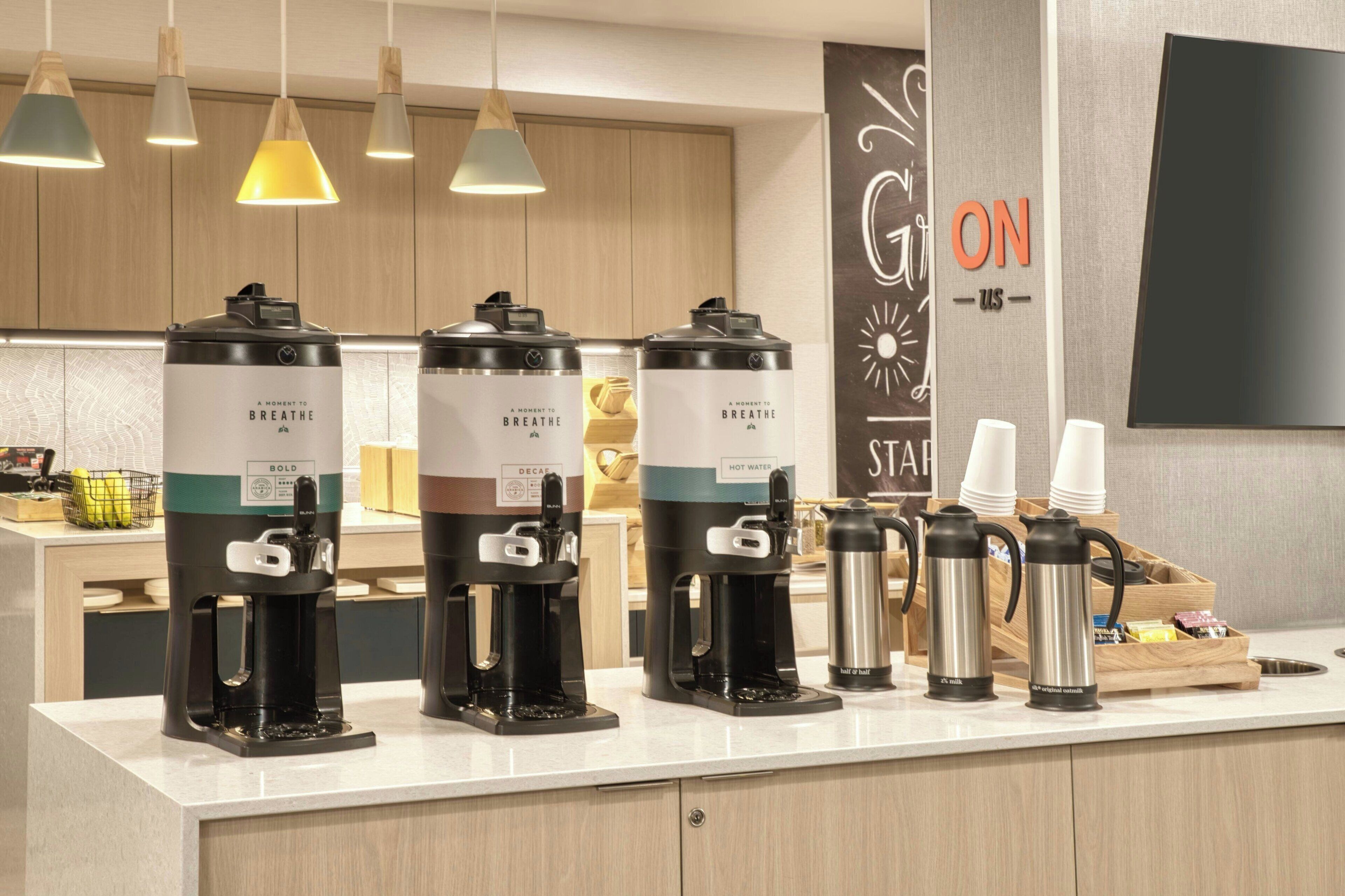 A coffee station with multiple coffee makers, a hot water dispenser, and a wall-mounted television.