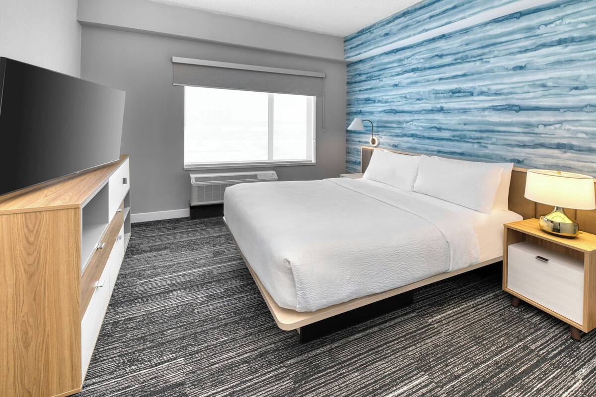 Una habitación de hotel con cama, mesita de noche, lámpara, televisor y ventana con persianas.