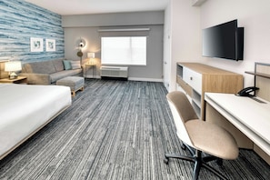 Habitación de hotel con cama, escritorio, silla, televisión de pantalla plana, ventana con persianas, aire acondicionado instalado en la pared y lámpara.