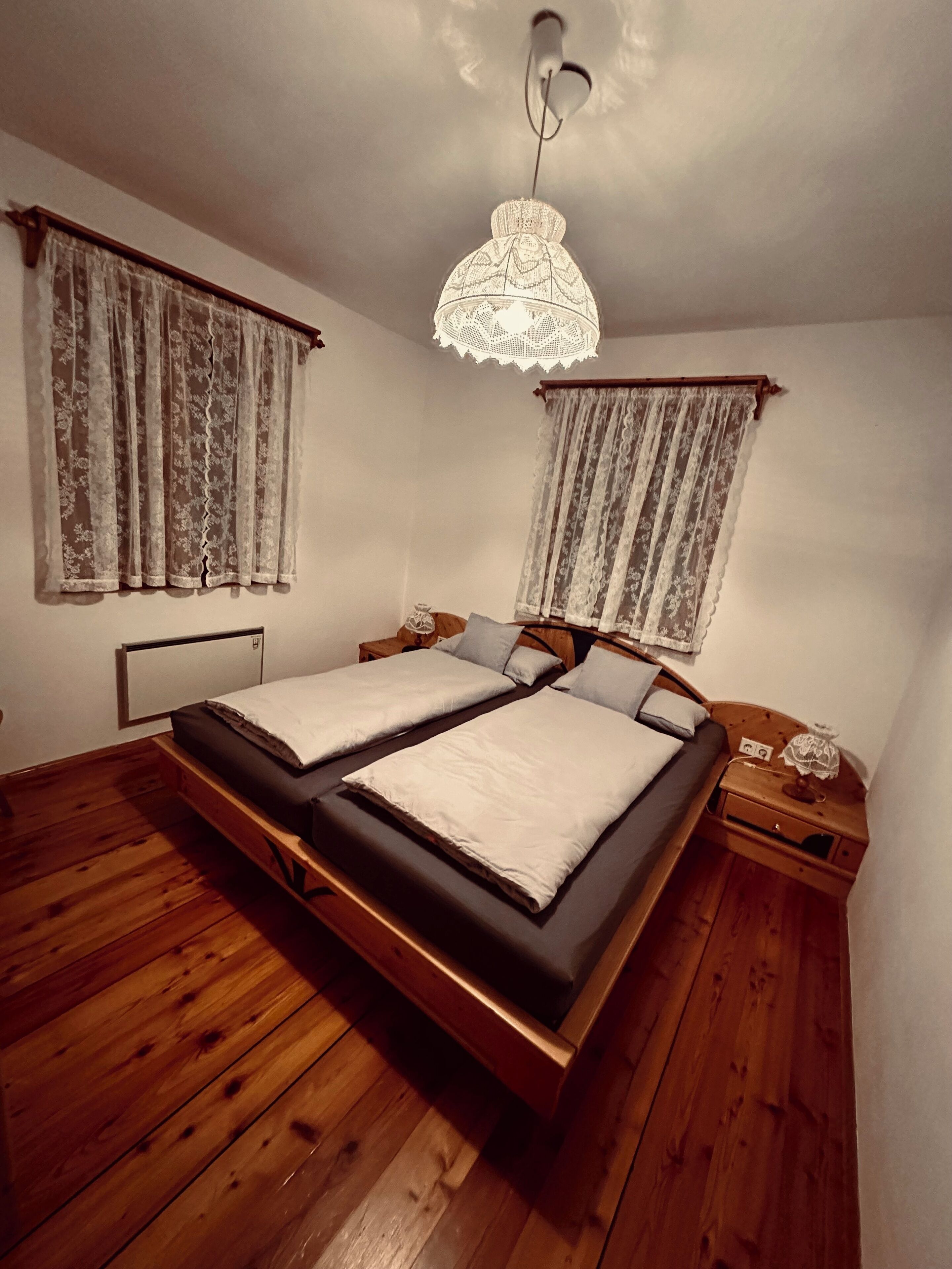 A room with a wooden floor, two beds with white linens, lace curtains, a wooden nightstand, and a hanging light fixture.