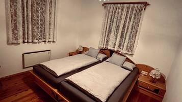 A room with a wooden floor, two beds with white linens, lace curtains, a wooden nightstand, and a hanging light fixture.