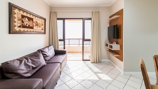 A modern living room with a sofa, a television mounted on the wall, and a balcony with curtains. - Fully Equipped Apartments in Meia Praia, 150m From the sea - Eoos (Itapema)
