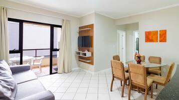 Una sala de estar moderna con un sofá, una mesa de comedor con sillas, un televisor montado en la pared y un balcón con vistas.