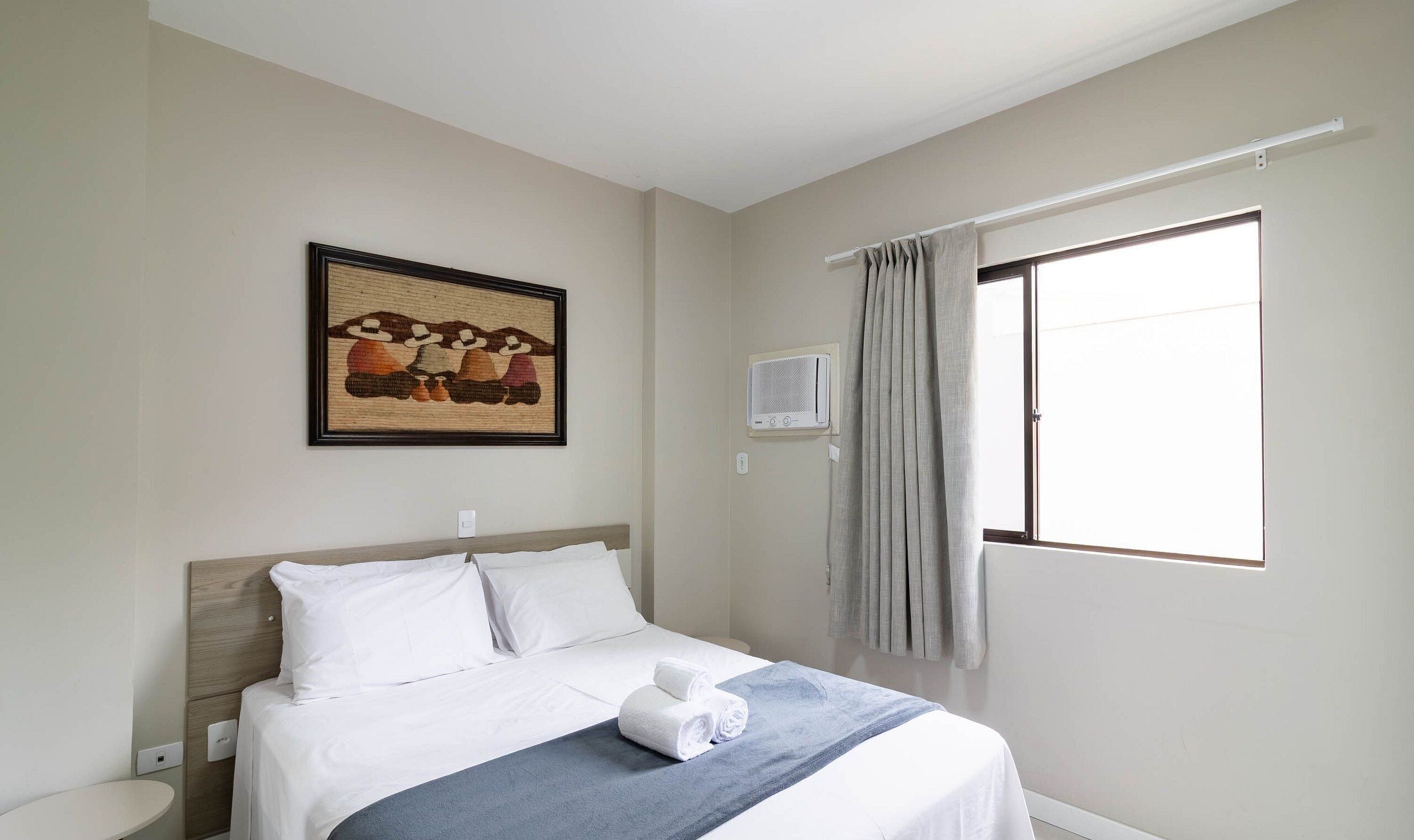 Quarto de hotel com cama, quadro na parede, aparelho de ar condicionado e janela com cortinas.