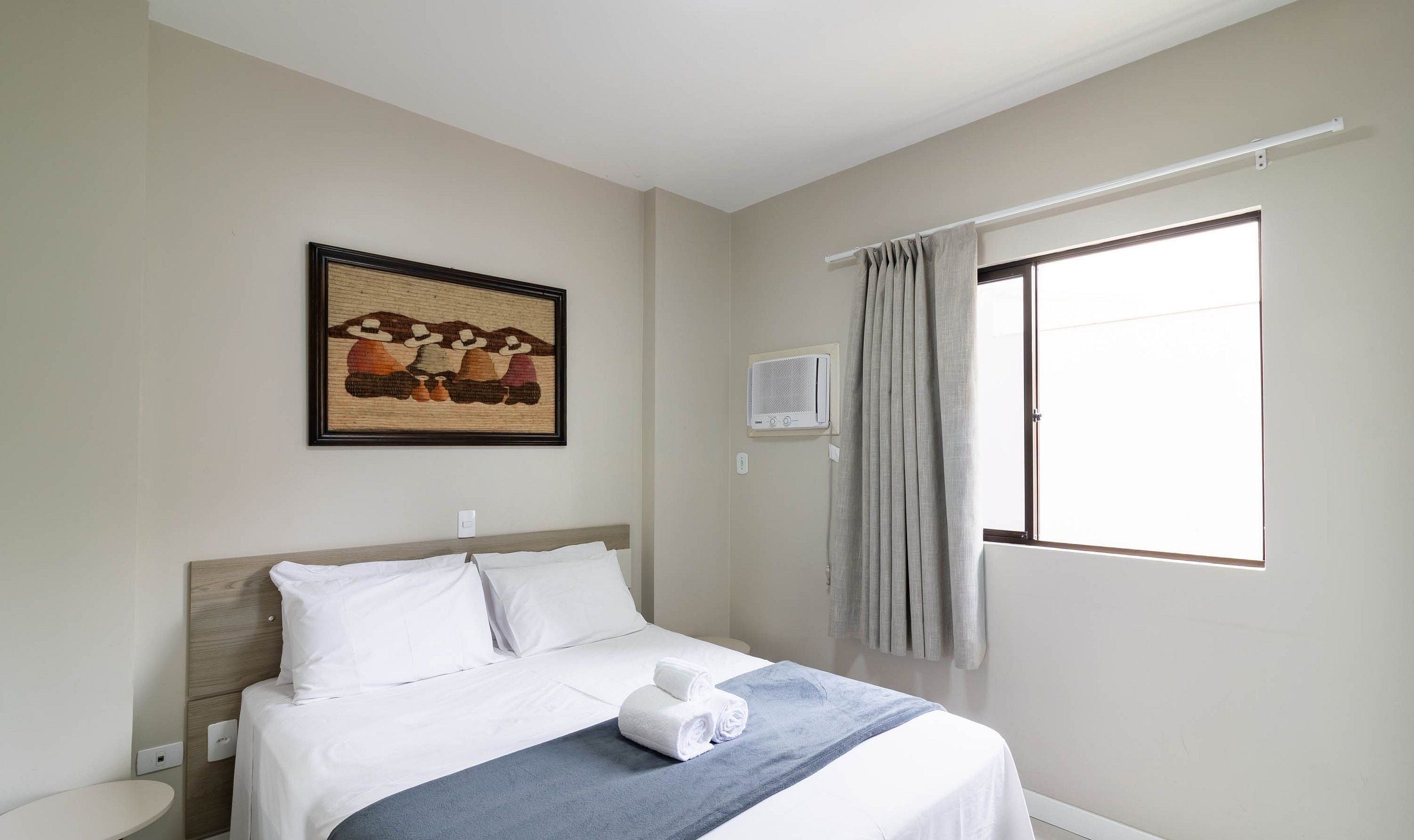 Une chambre d’hôtel avec un lit, un tableau au mur, un appareil de climatisation et une fenêtre avec des rideaux.