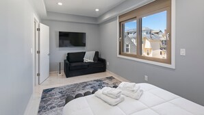 Une chambre moderne avec un grand lit, un canapé noir, un téléviseur fixé au mur et une fenêtre donnant sur les maisons voisines.
