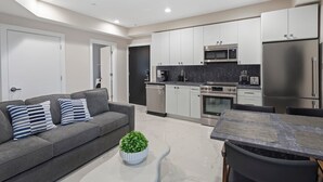 A modern kitchen with white cabinets, stainless steel appliances, and a grey sofa with striped pillows. - Ammos Luxury Living- Crete Suite (Ventnor City)