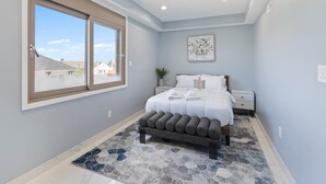 A bedroom with a large window, a bed with white linens, a gray bench, and a mosaic rug. - Ammos Luxury Living- Crete Suite (Ventnor City)