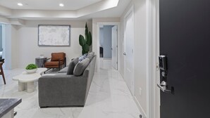 A modern living room with a grey sofa, a coffee table, and a brown armchair. There is a large abstract painting on the wall and a potted plant. - Ammos Luxury Living- Crete Suite (Ventnor City)