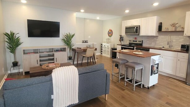 Een moderne keuken met witte kastjes, een magnetron en een spoelbak. Er is een eethoek met een tafel en stoelen, en een woonkamer met een bank en een televisie.