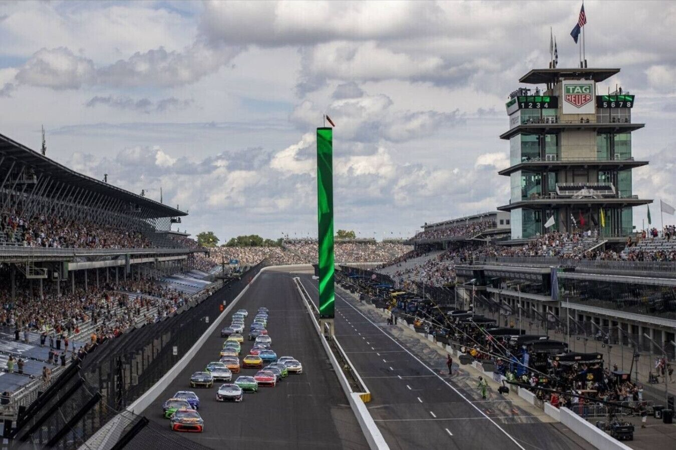A race track with multiple cars lined up, a green flag, and a control tower.