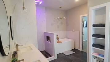 A modern bathroom with a large sink, a bathtub, and a walk-in shower. There is a vanity with a mirror and a shelf with towels.