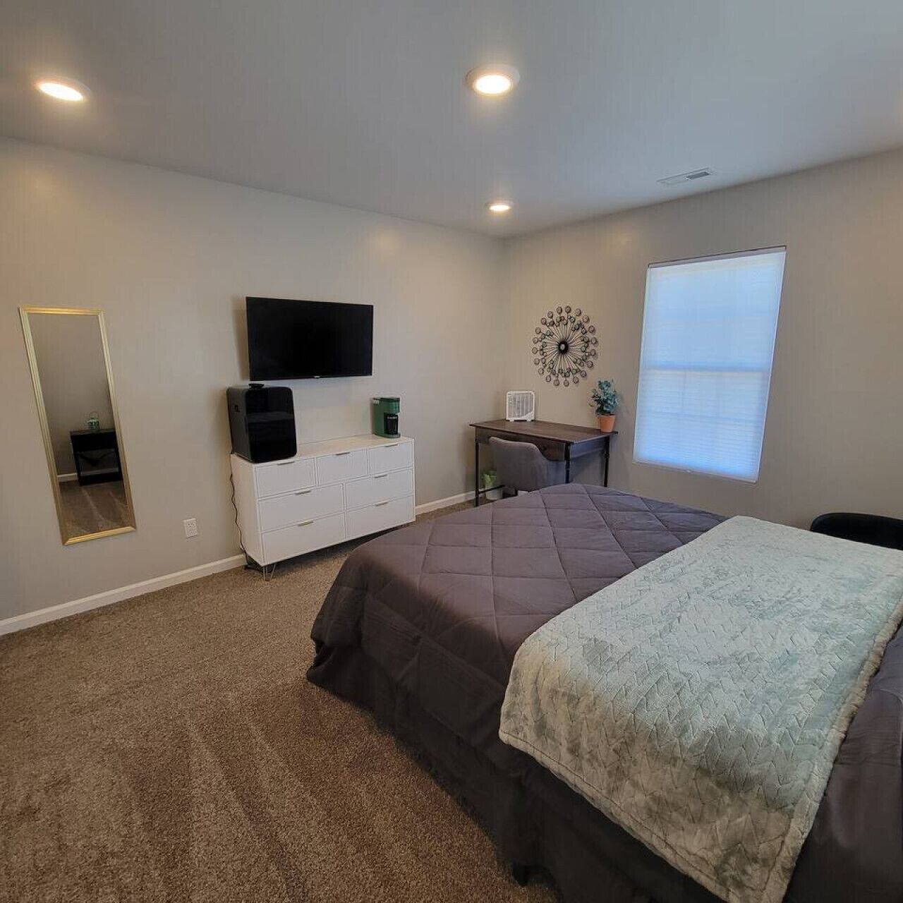 A bedroom with a bed, a dresser, a TV, a desk, and a window.