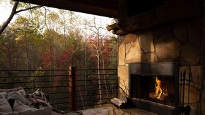 Une cheminée en pierre avec un feu de bois, un banc en bois et une vue sur les arbres d’automne.