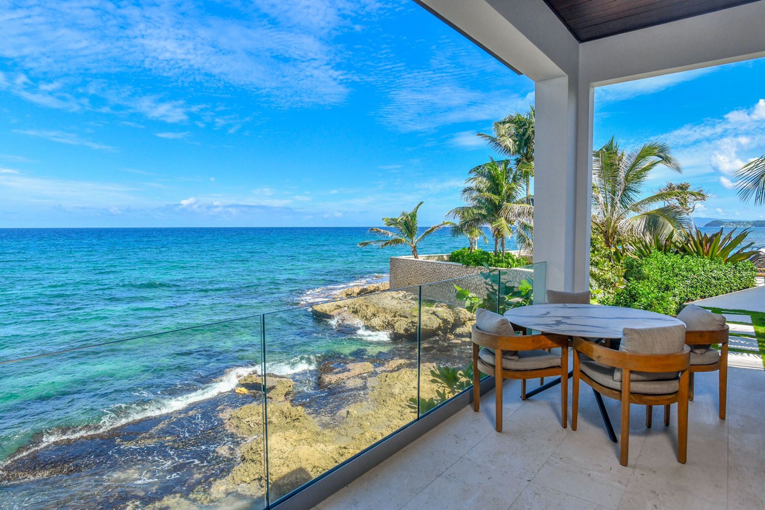 A balcony with a view of the ocean, a table with chairs, and palm trees.
