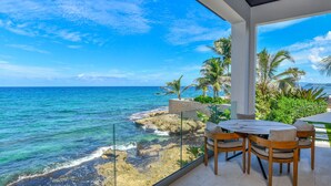 A balcony with a view of the ocean, a table with chairs, and palm trees. - Oceans 8 Villa 6BR (Sandy Bay)