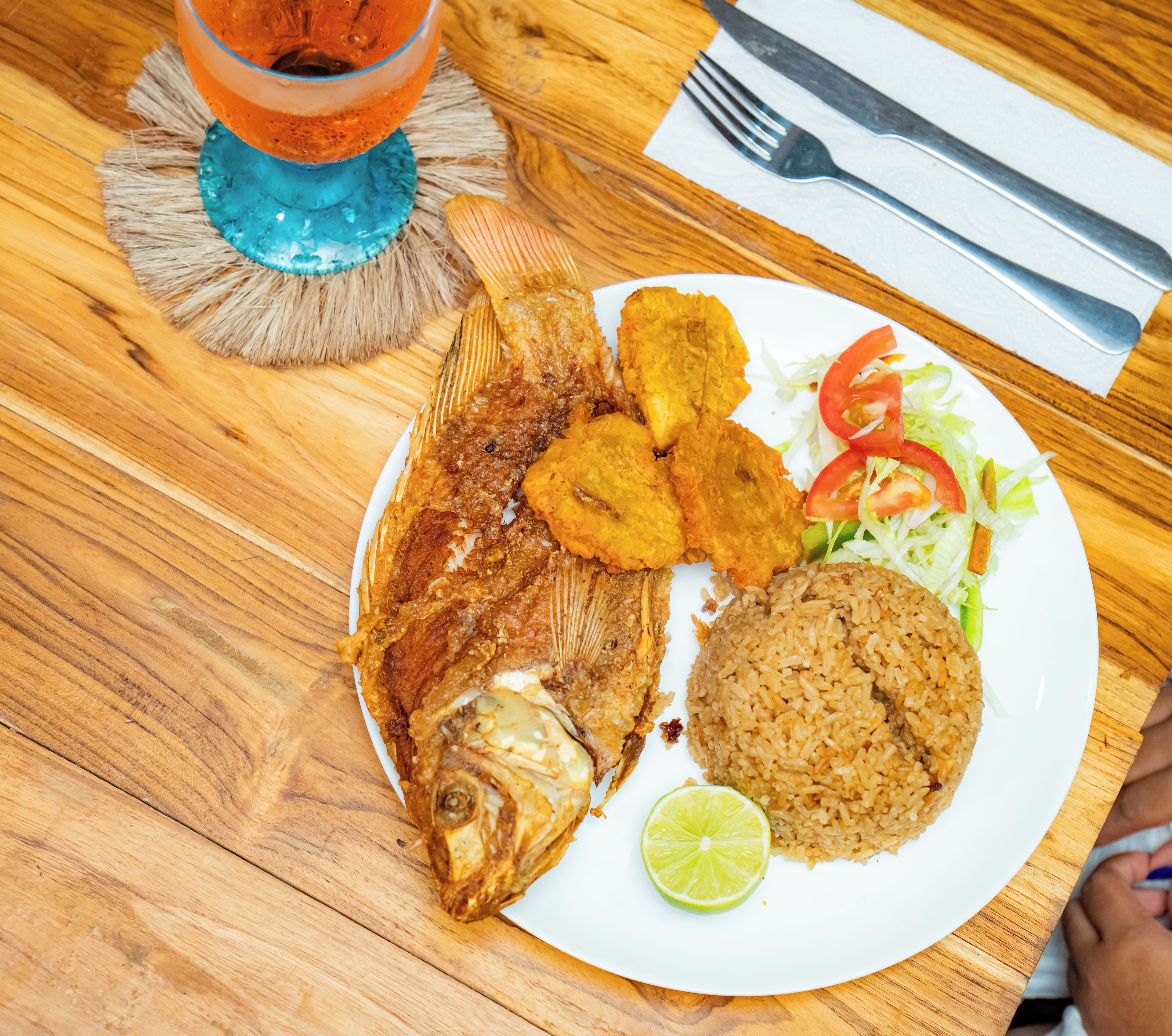 A plate with fried fish, fried plantains, rice, and a slice of lime on a wooden table.
