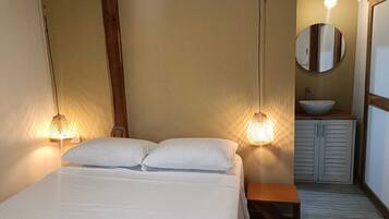 A bedroom with a bed, bedside lamps, a wooden bench, a round mirror, and a sink with a bowl.