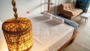 A bed with a white mattress and a wooden headboard, a hanging woven light fixture, and a beige sofa in the background. - Mar Barú Beach House (Cartagena)