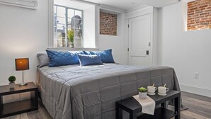 Une chambre moderne avec un grand lit, une table de chevet, une petite table avec deux tasses et une plante, et une fenêtre donnant sur l’extérieur.