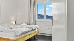 Una camera da letto con un letto di legno, biancheria bianca, un comodino con una lampada, una finestra vista montagne e un termosifone.