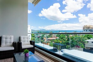 Un balcón con dos sillas blancas acolchadas, una mesa de vidrio y una planta en maceta. El balcón ofrece vistas a un paisaje urbano con edificios y áreas verdes.