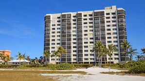 A high-rise hotel with multiple floors, surrounded by palm trees and a sandy area.