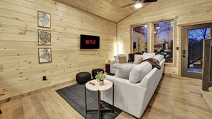 A modern living room with a wooden interior, a white sofa, a small round table with a plant, and a large TV screen displaying 'NETFLIX'. - Modern Cabin Retreat w/ Hot Tub, Fire Pit & Arcade near Pigeon Forge - Pets OK (Sevierville)