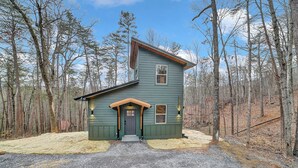 A two-story cabin with a green exterior, a front porch, and a gravel driveway surrounded by trees. - Modern Cabin Retreat w/ Hot Tub, Fire Pit & Arcade near Pigeon Forge - Pets OK (Sevierville)