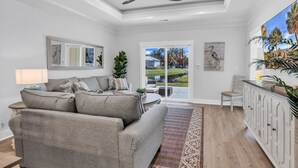 A modern living room with a grey sofa, a dining area with a white cabinet, and a large window with a view of a garden. - Luxury Pondside Retreat (Murrells Inlet)