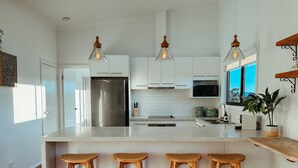 A modern kitchen with a large island, white cabinets, stainless steel appliances, and three wooden stools. - Snapper Hideaway - Pet Friendly (Merimbula)