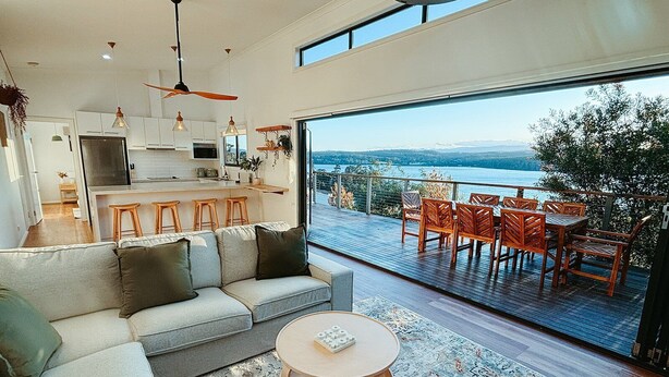 A modern living space with a kitchen, a sofa, a round coffee table, and a balcony with a view of the water. - Snapper Hideaway - Pet Friendly (Merimbula)