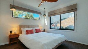 A bedroom with a large bed, two bedside tables, a ceiling fan, and a window with a view of the outdoors. - Snapper Hideaway - Pet Friendly (Merimbula)