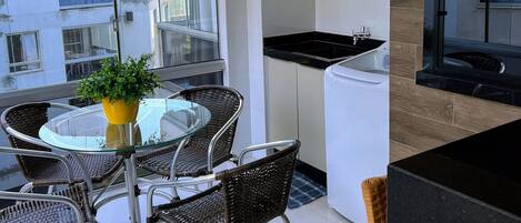 A compact balcony with a glass table and wicker chairs, a small potted plant, a compact kitchen with a sink and washing machine, and a view of the cityscape.