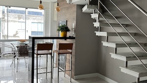 A modern kitchen with a glass dining table, two bar stools, a potted plant, and a staircase with a metal railing. - Apartamento Itapema (Itapema)
