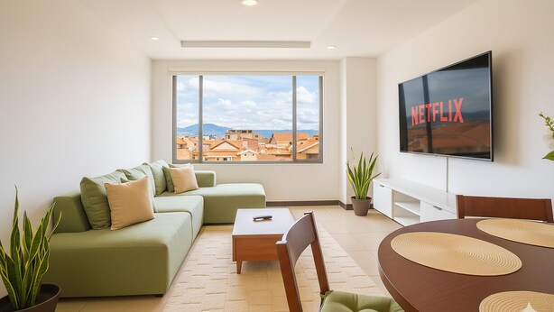 Una sala de estar moderna con un sofá verde, una mesa de centro de madera y una mesa de comedor con individuales. Una pantalla de televisor grande muestra el logotipo de Netflix.