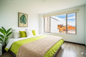 Ein Schlafzimmer mit einem großen Bett, Blick auf Gebäude durch ein Fenster und einer Zimmerpflanze.