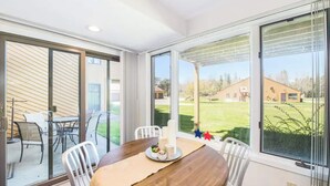 A dining area with a wooden table, white chairs, and a view of a grassy yard through large windows.