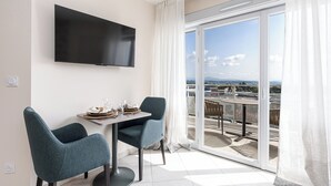 Een moderne kamer met een flatscreen televisie aan de muur, een eettafel met twee stoelen, en een glazen schuifdeur naar een balkon met uitzicht over de stad.