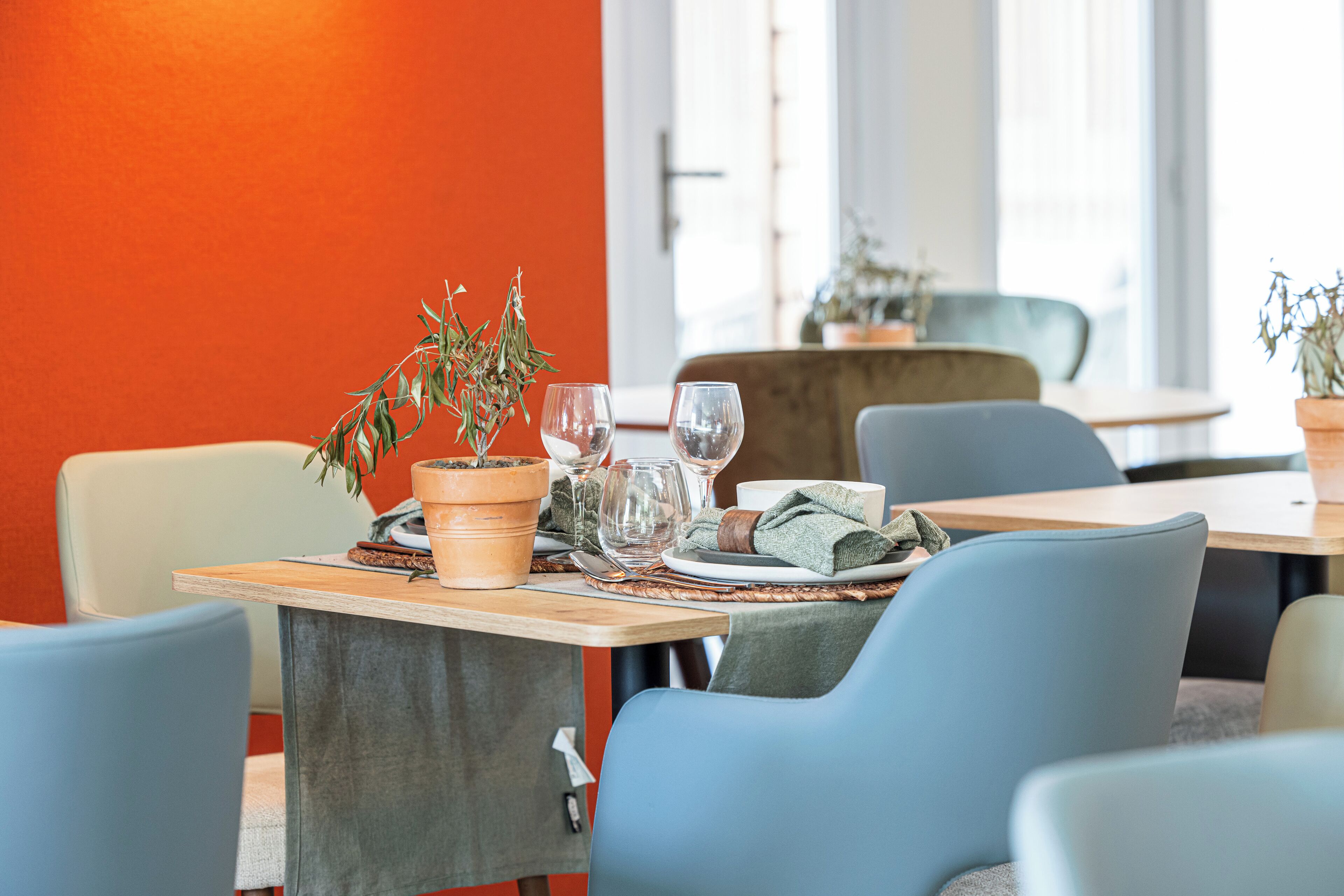 A dining area with a wooden table set for two, featuring a potted plant, wine glasses, and a cloth napkin.