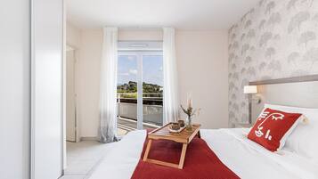 Une chambre d’hôtel avec un lit, une petite table avec un vase et une tasse, un plaid rouge et une vue sur l’extérieur par une grande fenêtre.