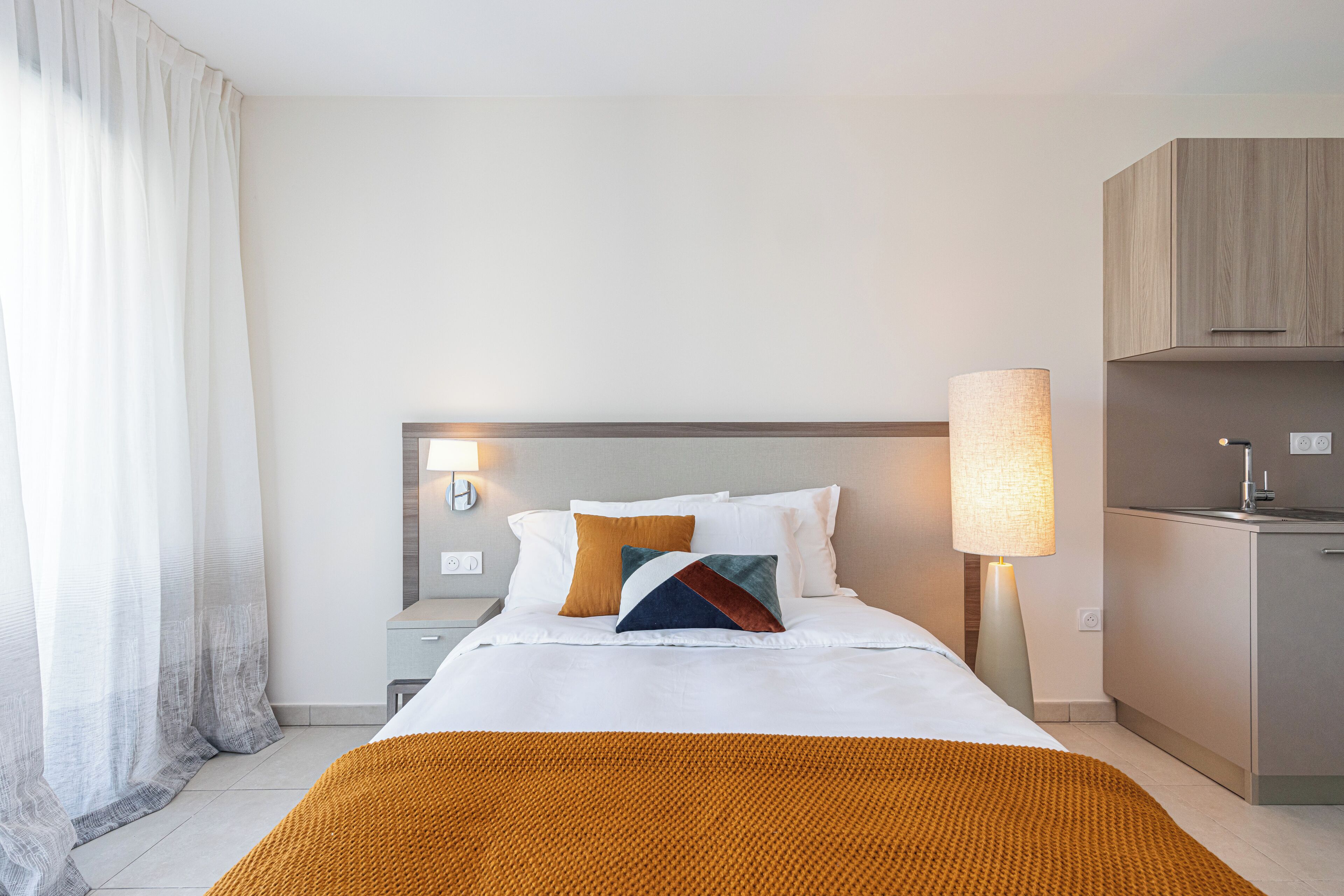 A hotel room with a bed, bedside lamps, a nightstand, a desk, and a kitchenette.